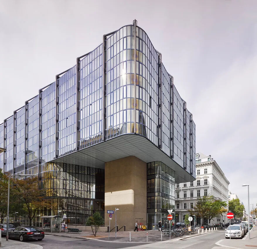 The Juridicum seen from the street corner: a curved glass-and-steel block whose upper floors cantilever out over a recessed, stone-clad ground floor, with the older houses of the Schottenbastei alongside.
