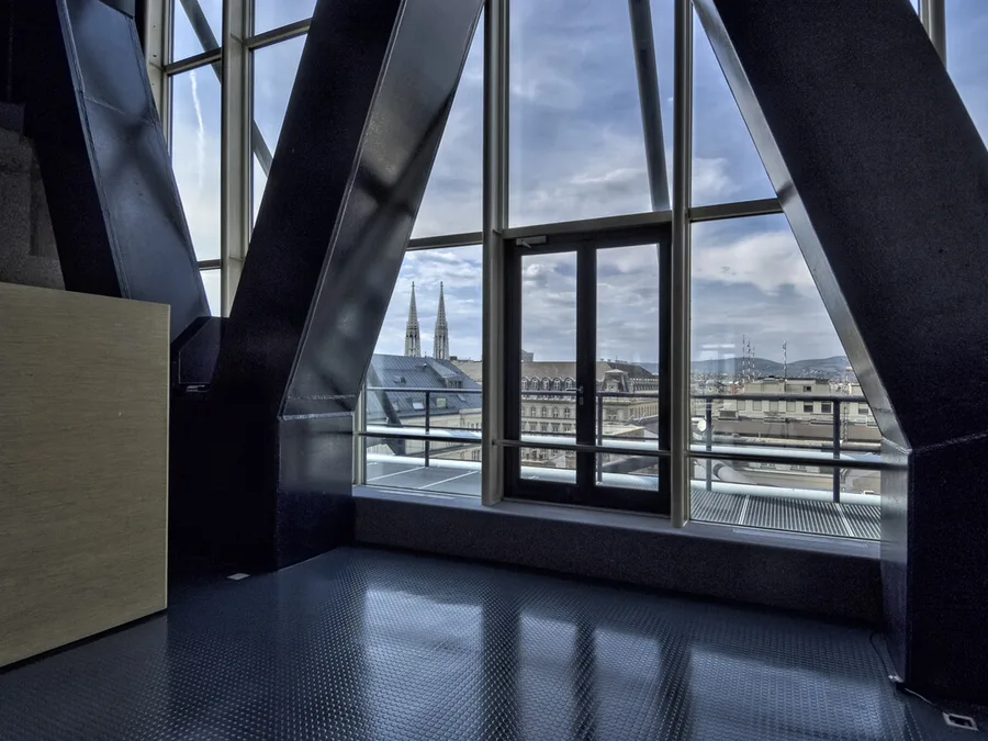 A room on the Juridicum's top floor, framed by dark steel trusses, looking through a glass wall over Vienna's rooftops to the twin spires of the Votivkirche.
